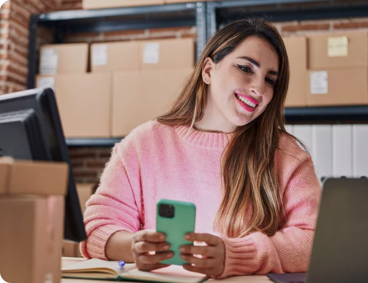 Lojista sorrindo com o celular na mão, entre caixas de pedidos